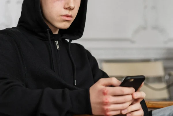 Nastolatek w czarnej bluzie z kapturem patrzy w smartfon, ilustrując wycofanie i lęk społeczny.