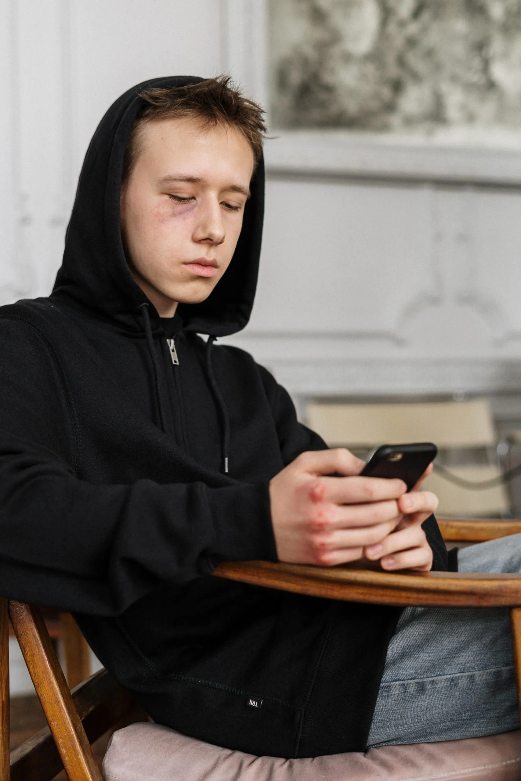 Nastolatek w czarnej bluzie z kapturem patrzy w smartfon, ilustrując wycofanie i lęk społeczny.